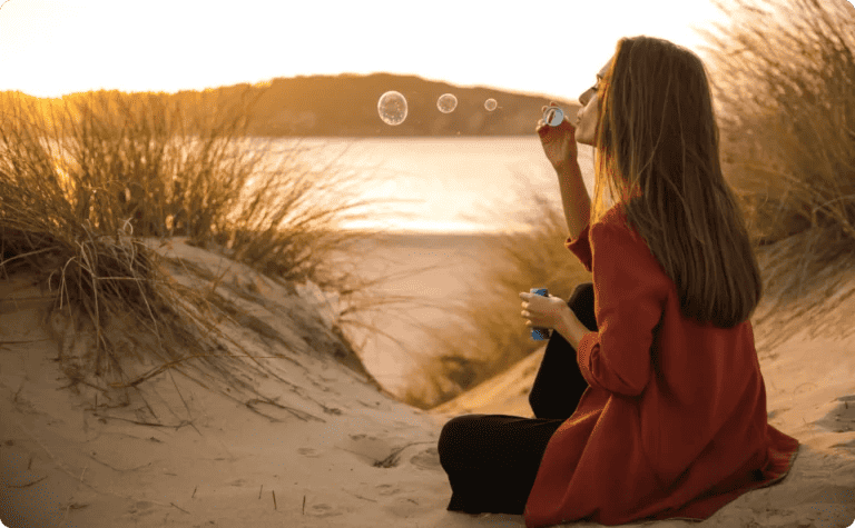 A woman sitting on a sand dune at sunset, blowing bubbles as a therapeutic exercise.