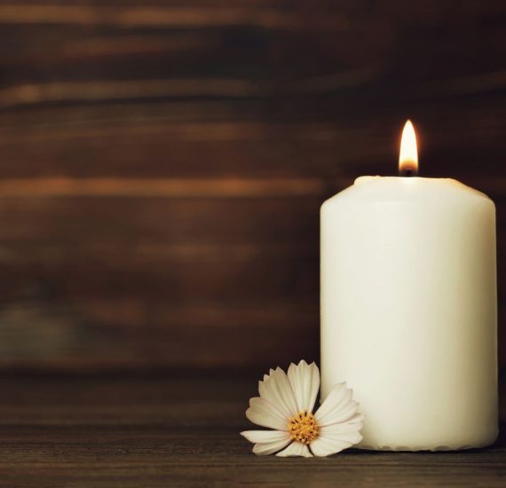 A lit white candle with a single white flower on a wooden background, symbolizing remembrance and reflection.